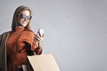 Laptop Stand - Smiling female in trendy wear and stylish sunglasses standing with paper shopping bags on background of gray wall and browsing mobile phone