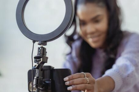 Smartphone Tripod - Cheerful young African American female blogger in stylish sweater smiling while setting up camera of smartphone attached to tripod with ring light before recording vlog