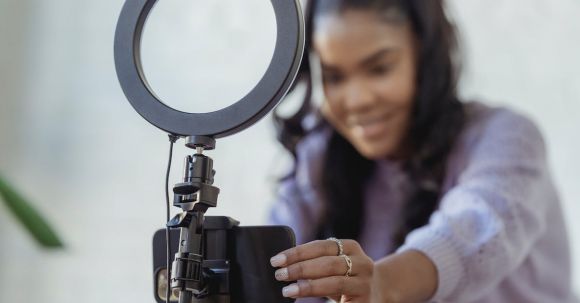 Smartphone Tripod - Cheerful young African American female blogger in stylish sweater smiling while setting up camera of smartphone attached to tripod with ring light before recording vlog