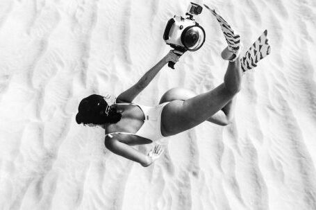 Underwater Cameras - Black and white faceless slim female wearing swimsuit and flippers snorkeling above sandy sea bottom in transparent waters and carrying camera in waterproof cover