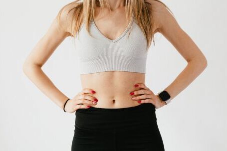 Sync Wearable - Faceless slim anonymous blond female in sports bra and black leggings in wearable bracelet showing perfect belly on white background while standing with hands on waist