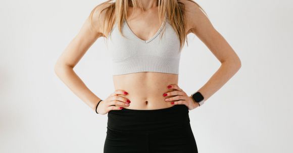 Sync Wearable - Faceless slim anonymous blond female in sports bra and black leggings in wearable bracelet showing perfect belly on white background while standing with hands on waist